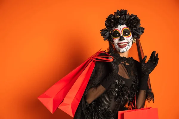 Улыбающаяся женщина в черном костюме и жутком макияже на Хэллоуин, держащая сумки с покупками и размахивая рукой на оранжевом фоне — стоковое фото