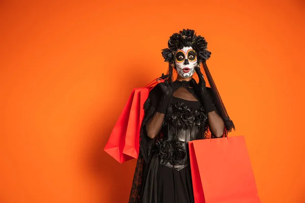 Überraschte Frau in gruseligem Halloween-Make-up und schwarzem Kostüm mit Einkaufstüten auf orangefarbenem Hintergrund — Stockfoto