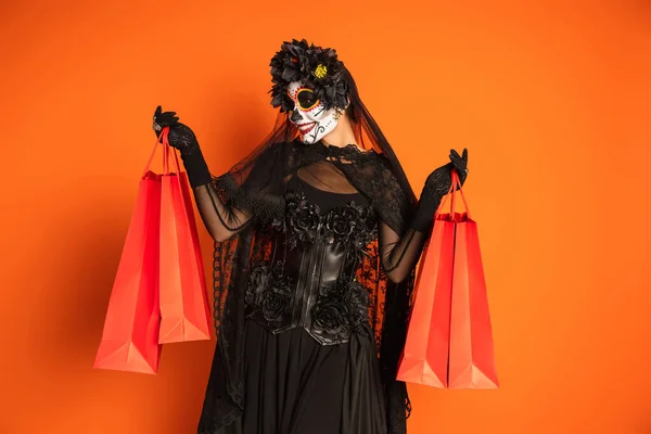 Alegre mujer en santa muerte maquillaje y negro traje celebración bolsas aisladas en naranja - foto de stock