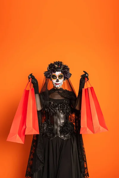 Femme en maquillage catrina et costume noir effrayant avec couronne montrant des sacs à provisions isolés sur orange — Photo de stock