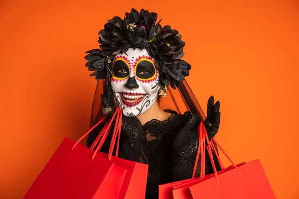 Mujer alegre en corona negra y el cráneo de azúcar de Halloween maquillaje mirando a la cámara cerca de bolsas de compras aisladas en naranja - foto de stock