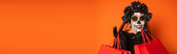 Glückliche Frau in schwarzem Kranz und gruseligem Halloween-Make-up, die in die Kamera neben Einkaufstüten schaut, vereinzelt auf orangefarbenem Banner — Stockfoto