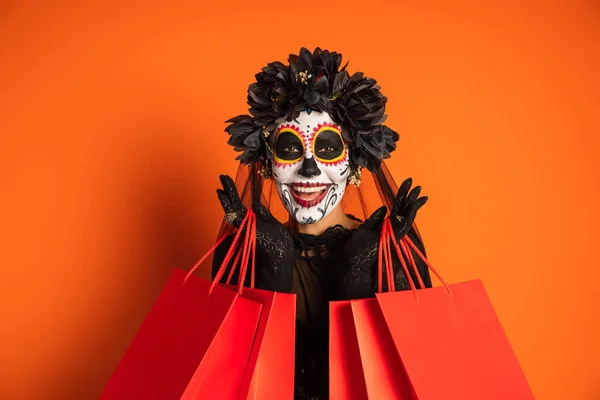 Donna sorridente in costume di Halloween nero e trucco teschio di zucchero che tiene borse della spesa e guardando la fotocamera su sfondo arancione — Foto stock