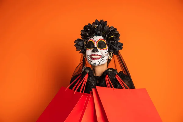 Donna felice con gli occhi chiusi e trucco catrina spettrale che tiene borse della spesa isolate su arancione — Foto stock