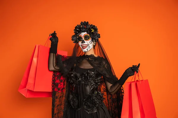 Mulher feliz em traje de bruxa preta e maquiagem halloween segurando sacos de compras e olhando para longe isolado em laranja — Fotografia de Stock