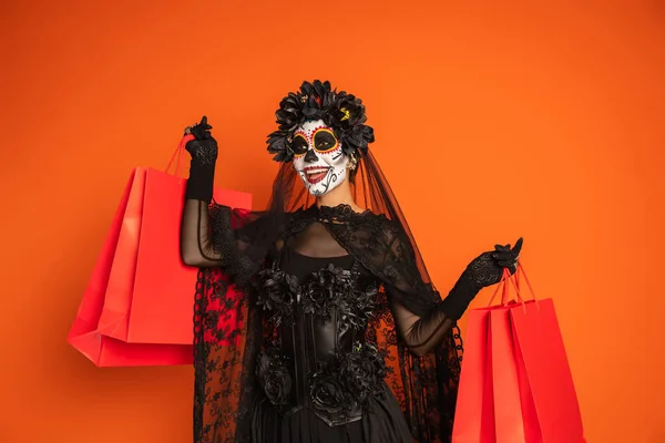 Happy woman in black witch costume and halloween makeup holding shopping bags and looking away isolated on orange — Stock Photo