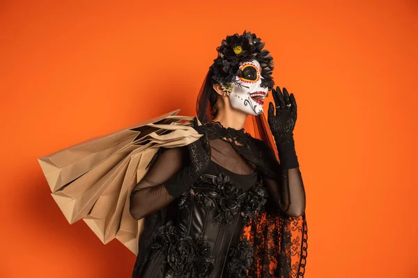 Donna eccitata in costume nero Santa Muerte e trucco tenendo borse della spesa e urlando a qualcuno isolato su arancione — Foto stock