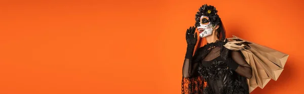 Donna felice in costume nero raccapricciante e trucco teschio di zucchero che tiene borse della spesa e urla a qualcuno isolato su arancione, banner — Foto stock