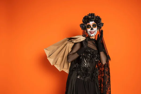 Femme heureuse au Mexique jour de maquillage mort et costume noir debout avec des sacs à provisions sur fond orange — Photo de stock