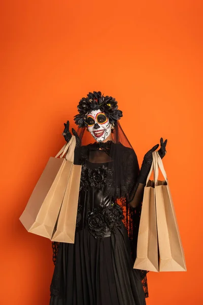 Mujer en día mexicano de disfraz muerto y maquillaje catrina sonriendo a la cámara con bolsas de compras aisladas en naranja - foto de stock