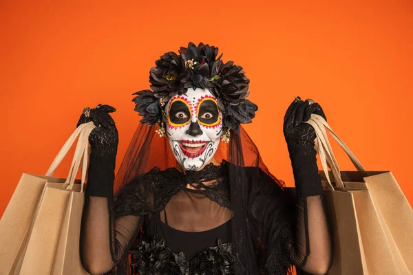 Mulher espantada em traje de Halloween preto e maquiagem assustadora segurando sacos de compras isolados em laranja — Fotografia de Stock