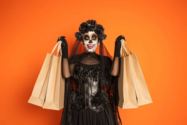 Mujer excitada en traje de halloween negro con corona y maquillaje aterrador sosteniendo bolsas aisladas en naranja - foto de stock