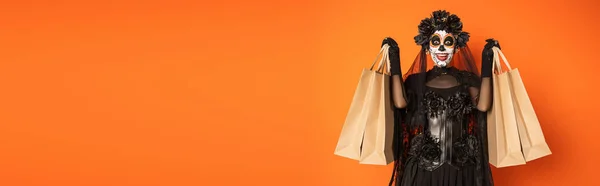 Mulher espantada no dia mexicano de traje morto e maquiagem mostrando sacos de compras isolados em laranja, banner — Fotografia de Stock
