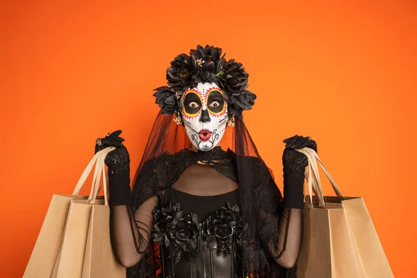 Mujer sorprendida en el día tradicional de disfraz muerto y maquillaje catrina mostrando bolsas de compras aisladas en naranja - foto de stock