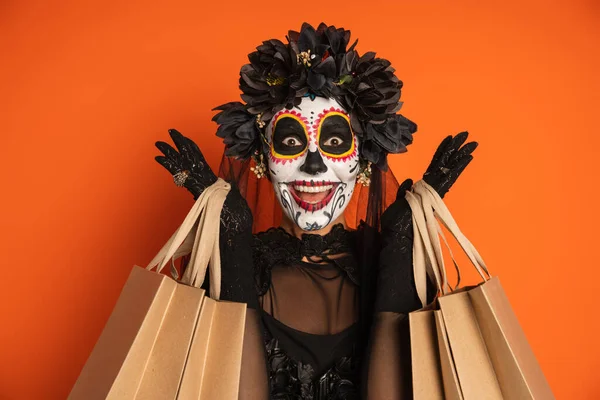 Mujer excitada en espeluznante maquillaje de halloween y corona negra sosteniendo bolsas de compras y mirando a la cámara aislada en naranja - foto de stock