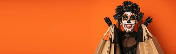 Mujer emocionada en el maquillaje de Halloween espeluznante y corona negra sosteniendo bolsas de compras aisladas en naranja, pancarta - foto de stock