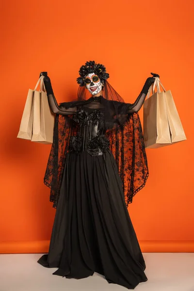 Longitud completa de la mujer alegre en vestido de bruja negro y maquillaje de Halloween mostrando bolsas de compras sobre fondo naranja - foto de stock