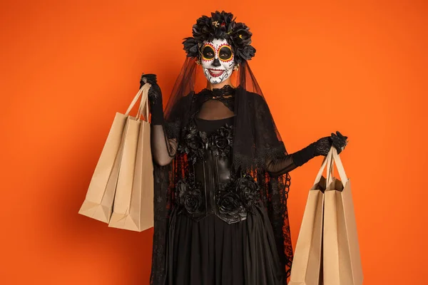 Gioiosa donna in tradizionale trucco Santa Muerte e costume nero in piedi con borse della spesa isolate su arancione — Foto stock