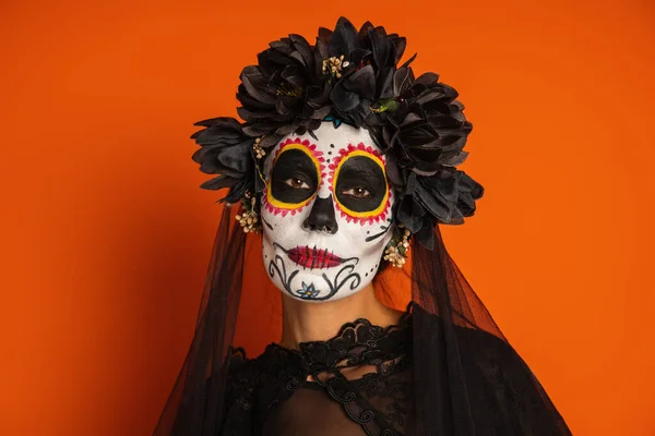 Retrato de la mujer en corona negra y el cráneo de azúcar de Halloween maquillaje mirando a la cámara aislada en naranja - foto de stock