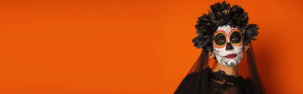 Портрет женщины в мексиканский день мертвого макияжа и черного костюма, смотрящей в камеру изолированной на оранжевый, баннер — стоковое фото