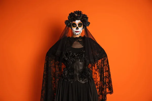 Женщина в традиционной санта муэрте макияж и костюм с черной кружевной вуаль стоя с руками за спиной изолированы на оранжевый — стоковое фото