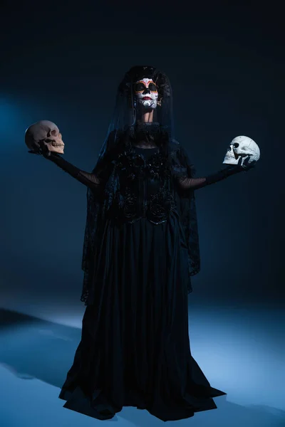 Donna in trucco spettrale e vestito da strega in piedi con gli occhi chiusi e teschi su sfondo scuro con luce blu — Foto stock
