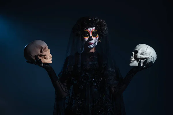 Vue de face de la femme dans le maquillage d'Halloween et costume noir debout avec des crânes sur fond bleu foncé — Photo de stock
