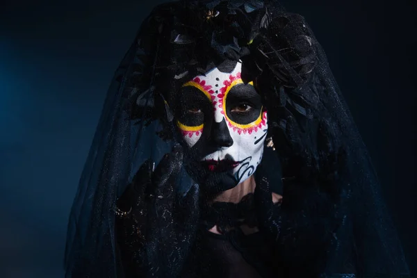 Woman in sugar skull halloween makeup gesturing near black veil on dark blue background — Stock Photo