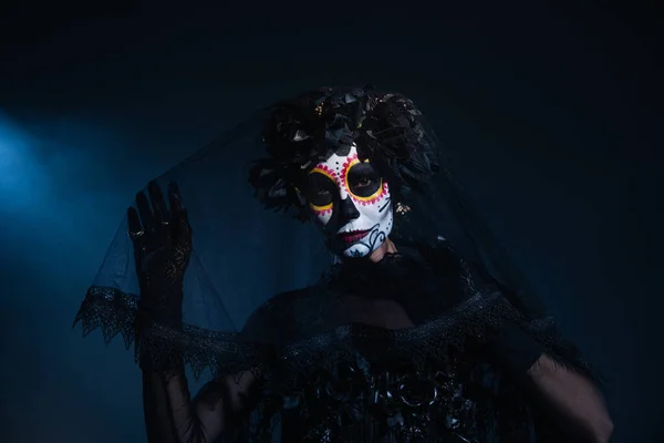 Frau in traditionellem santa muerte Make-up berührt schwarzen Schleier und blickt in die Kamera auf dunklem Hintergrund mit blauem Licht — Stockfoto