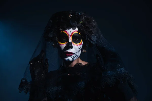 Woman in mexican day of dead costume and catrina makeup looking at camera under black lace veil on dark blue background — Stock Photo