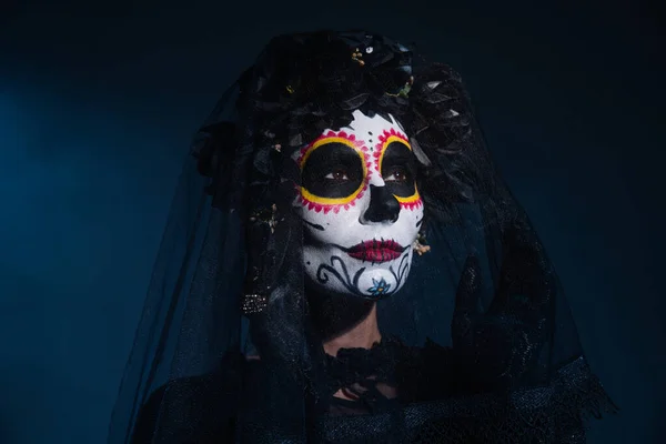 Femme dans le traditionnel mexicain catrina maquillage toucher voile et regarder loin sur fond bleu foncé — Photo de stock