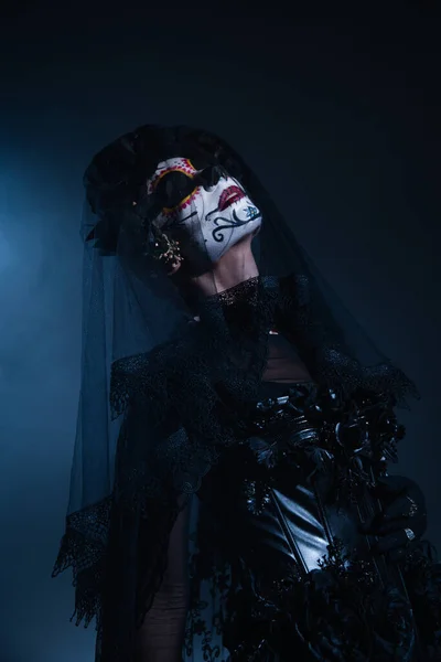 Mujer en espeluznante maquillaje de santa muerte y traje negro posando con los ojos cerrados sobre fondo negro y azul - foto de stock