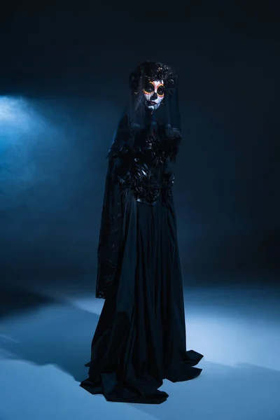 Longitud completa de la mujer en vestido de bruja negro y maquillaje de Halloween espeluznante sobre fondo oscuro con luz azul, bandera - foto de stock