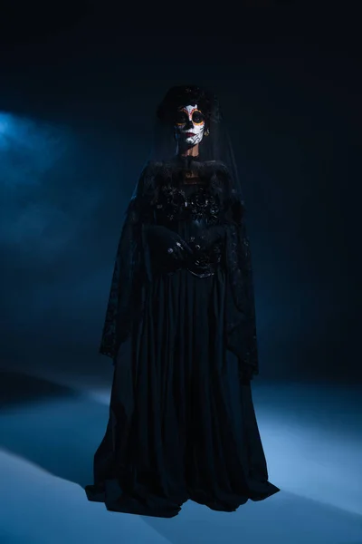 Full length of woman in traditional santa muerte makeup and black dress standing in blue light on dark background — Stock Photo