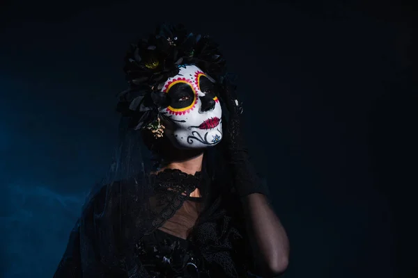 Frau mit Totenkopf-Halloween-Make-up hält Hand in Gesichtsnähe auf schwarzem und blauem Hintergrund — Stockfoto