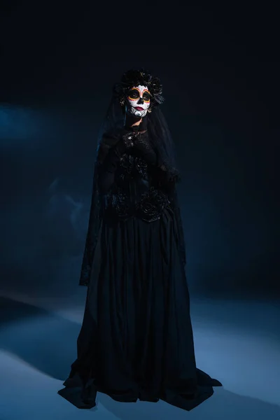 Volle Länge der Frau in schwarzem Hexenkleid und Halloween-Make-up auf dunklem Hintergrund mit blauem Licht — Stockfoto