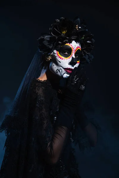 Woman in traditional mexican day of dead makeup and costume standing with praying hands on dark blue background — Stock Photo