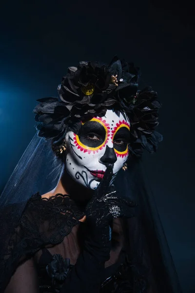 Mujer en el maquillaje de Halloween miedo y corona negra mostrando signo de silencio sobre fondo azul oscuro - foto de stock