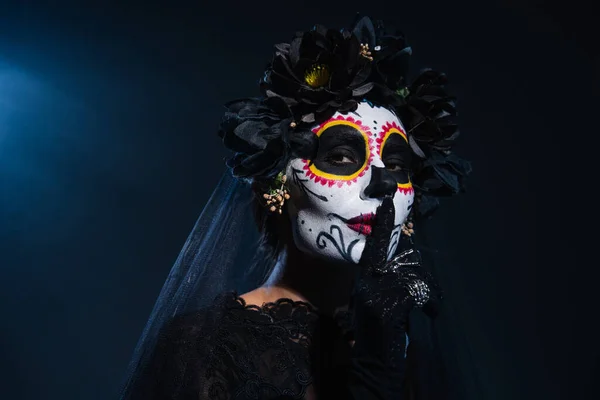 Mujer en espeluznante maquillaje de halloween y corona negra con velo mostrando signo de silencio sobre fondo azul oscuro - foto de stock