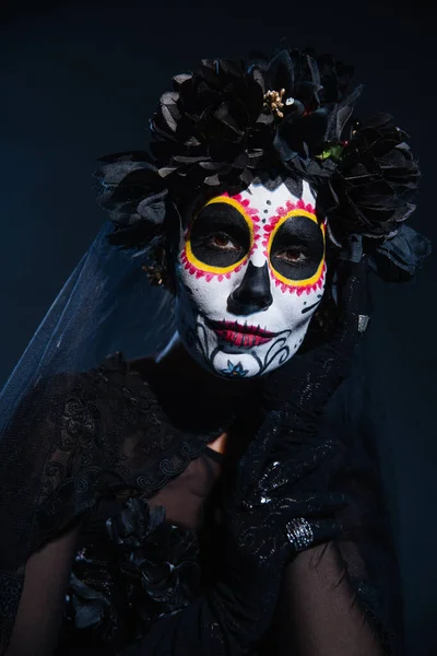 Portrait de femme en santa muerte maquillage traditionnel et couronne noire regardant la caméra sur fond bleu foncé — Photo de stock