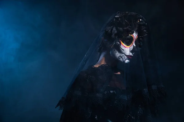 Femme dans le maquillage du crâne de sucre et costume noir d'Halloween debout avec les yeux fermés sur fond sombre avec de la fumée bleue — Photo de stock