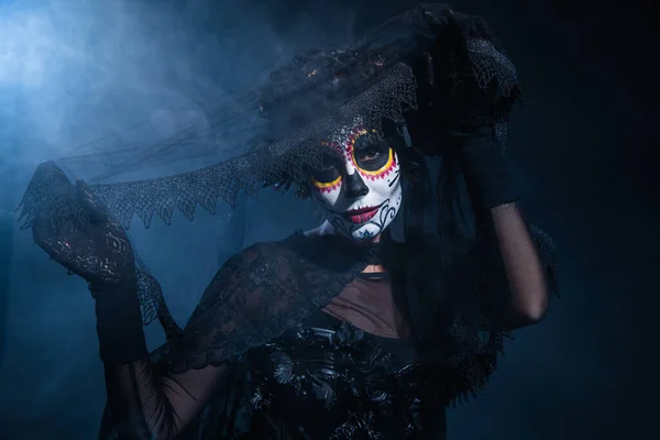 Femme dans le maquillage effrayant halloween tenant voile de dentelle noire et regardant la caméra sur fond sombre avec de la fumée bleue — Photo de stock
