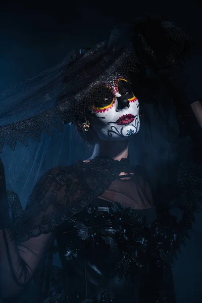 Mujer en traje de Halloween y maquillaje espeluznante posando con velo de encaje negro sobre fondo azul oscuro - foto de stock