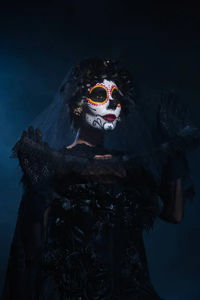 Woman in witch costume and sugar skull makeup holding lace veil on dark blue background — Stock Photo