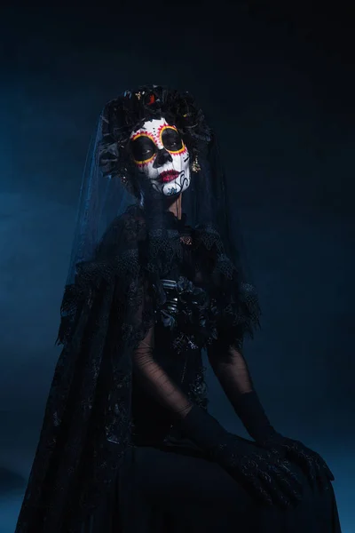 Femme en costume d'Halloween sorcière et maquillage du crâne de sucre assis sur fond bleu foncé — Photo de stock