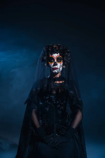 Frontansicht einer Frau im schwarzen Hexenkostüm und gruseligen Halloween-Make-up auf dunklem Hintergrund mit blauem Nebel — Stockfoto