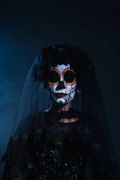 Donna in inquietante trucco di Halloween e ghirlanda nera con velo in posa con gli occhi chiusi su sfondo blu scuro — Foto stock