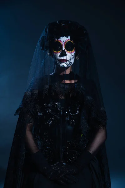 Mujer en tradicional mexicana santa muerte maquillaje y traje mirando a la cámara sobre fondo azul oscuro - foto de stock