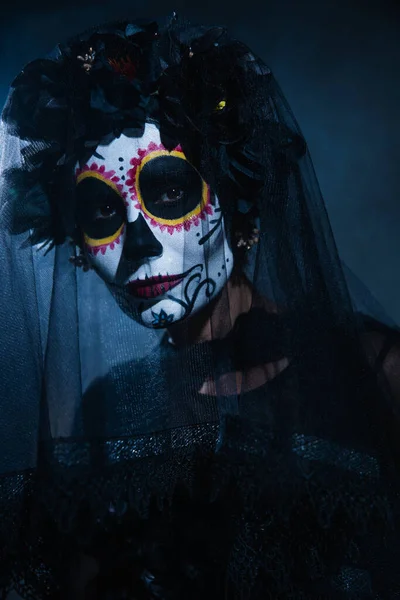 Portrait of woman in sugar skull halloween makeup looking at camera under black veil on dark background with blue light — Stock Photo
