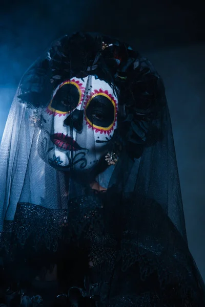 Woman in sugar skull halloween makeup and black wreath looking at camera under veil on dark background with blue light — Stock Photo